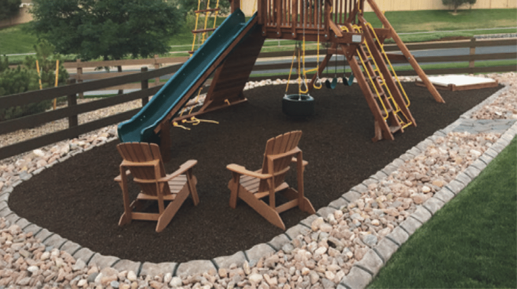 A backyard playground with a slide, swings, and two wooden chairs on mulch, bordered by rocks and grass, showcases expert execution in landscape design and concrete curbing installation.