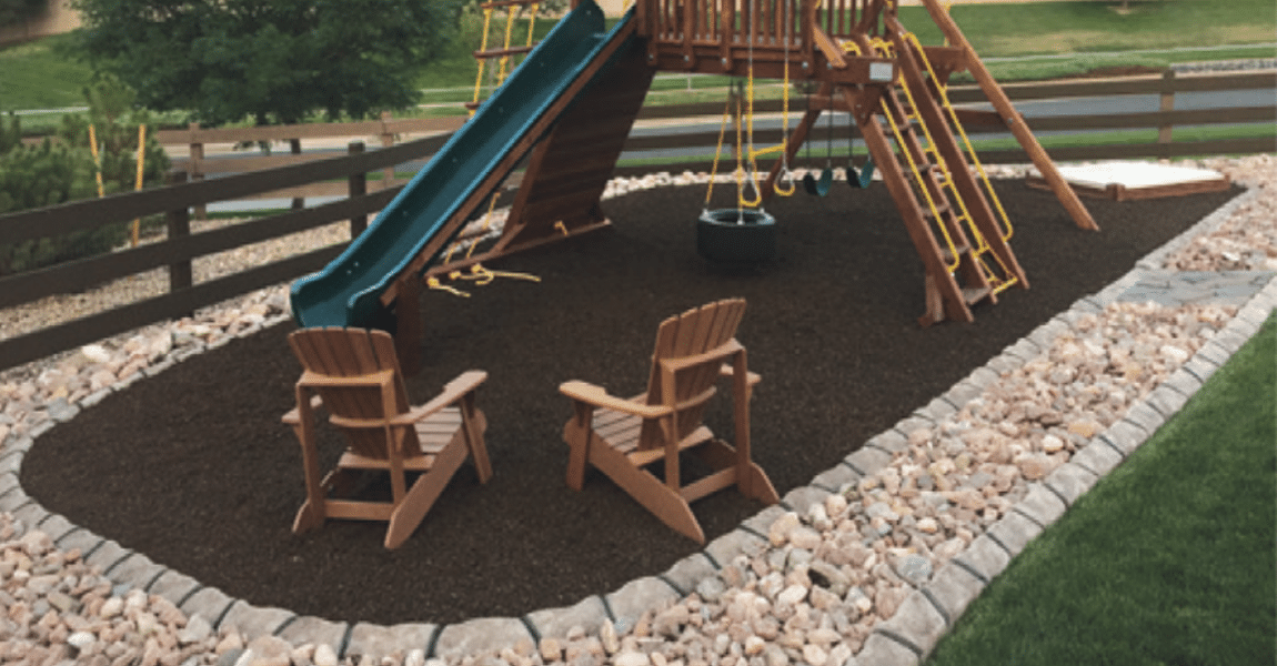 A backyard playground with a slide, swings, and two wooden chairs on mulch, bordered by rocks and grass, showcases expert execution in landscape design and concrete curbing installation.