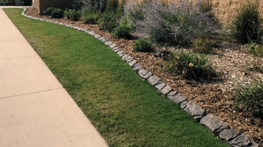 A sidewalk runs beside a neatly edged lawn and a landscaped garden bed with mulch, ornamental grasses, flowering shrubs, and concrete curbing that highlights the landscape’s professional installation process.