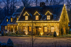 A stone house with snow on the roof is decorated with yellow string lights at dusk, its illuminated windows creating a warm gallery effect. Leafless trees in the background showcase the cozy winter scene.