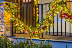 A large, decorated Christmas wreath with lights, ornaments, and ribbon hangs on a porch railing, creating a festive display that could brighten any art gallery or seasonal exhibitions at Showcase Gallery.