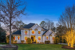 Two-story house in Showcase Gallery style, decorated for the holidays with wreaths and red bows on windows, and yellow string lights outlining trees and bushes in the yard at dusk.