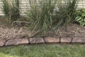 Showcase Gallery: Brown stone landscape edging separates a garden bed with tall grasses and mulch from a grassy lawn, set against a cream-colored siding wall in the background.