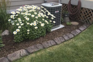 A flower bed with white daisies is next to an outdoor air conditioning unit and a garden hose, bordered by stones and mulch, beside a house and a lawn—an inviting scene worthy of any Showcase Gallery.