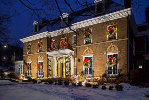 A large brick house decorated with garlands, red bows, and wreaths for the holidays, illuminated at night with some snow on the ground—like a festive showcase gallery celebrating winter’s charm.