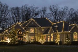 A large house is decorated with white holiday lights and wreaths on doors and windows, with trees and bushes in the yard also covered in lights, creating a festive Showcase Gallery of sparkling elegance.