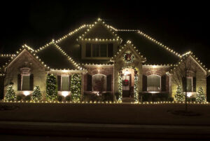 Single-story house showcased with white Christmas lights outlining the roof and bushes at night. Wreaths hang on the front door and above the entrance, creating a festive gallery of holiday cheer.