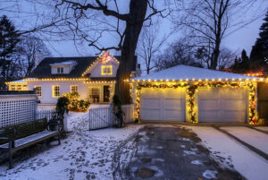 A house and garage are decorated with white and yellow holiday lights at dusk, surrounded by light snow on the ground and bare trees—a festive scene worthy of a Showcase Gallery.