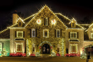 A large stone house decorated with Christmas lights around the roof, windows, and bushes, glowing brightly at night like an enchanting Showcase Gallery exhibition.