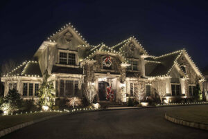 A large house decorated with white string lights and a wreath for the holidays becomes a festive showcase, with a figure dressed as Santa Claus standing at the front door at night.