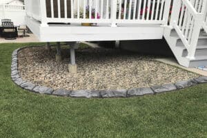 A section of yard under a raised deck creates a showcase gallery effect with a border of gray stones, filled with small rocks and surrounded by green grass.