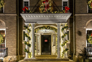 Front entrance of a brick house decorated with garlands, lights, and red bows for the holidays; a lit