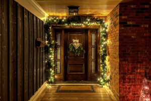A wooden front door decorated with a wreath and surrounded by garland with white string lights creates a stunning nighttime showcase, set in a porch with wood and brick walls—perfect for any gallery of inviting entrances.