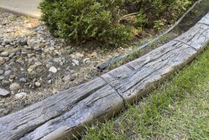 A close-up of a concrete garden edging that resembles wood, separating green grass from a rocky area with shrubs in the background—perfect for a Showcase Gallery of creative landscaping solutions.