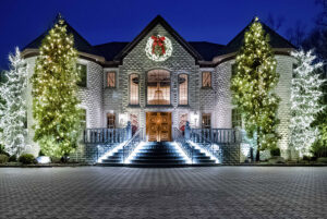 Large, two-story house decorated with white Christmas lights and a wreath above the entrance, flanked by illuminated trees at night, creates an exhibition-like ambiance reminiscent of an art gallery showcase.