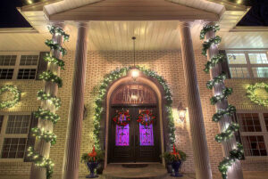 Front entrance of a house decorated with garlands and white string lights for the holidays, featuring wreaths and two planters by double doors at night—a true Showcase Gallery of festive charm.