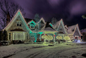 A large house with stone exterior is decorated with white and green holiday lights, creating a festive Showcase Gallery. Snow covers the ground and trees, and the sky is dark and cloudy.