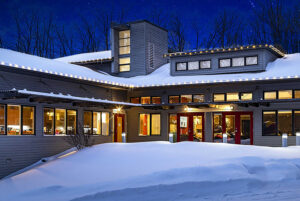 Modern house with large windows and string lights, surrounded by snow at night with trees in the background—resembling an exhibition at a Showcase Gallery, blending modern architecture with serene winter beauty.