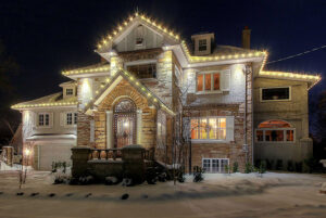 Large stone house at night with snow on the ground, lit by white string lights outlining the roof and architectural features—a true Showcase Gallery of winter elegance.