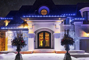 A large house with snow on the ground, illuminated at night and decorated with white string lights and holiday wreaths on the front doors—a true showcase of festive charm.