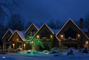 Large house with snow-covered roof is decorated with white and green holiday lights at night, surrounded by snowy trees and lawn—a true winter Showcase Gallery.