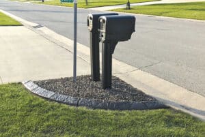 Two black mailboxes stand side by side on a gravel patch next to a sidewalk and street in a suburban neighborhood, resembling a quiet Showcase Gallery of everyday life.