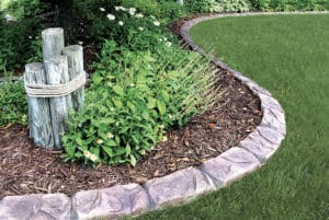 Curved stone garden edging separates a mulched flower bed with green plants and white flowers from a neatly trimmed lawn, creating a showcase gallery of natural beauty; wooden posts tied with rope are also visible.