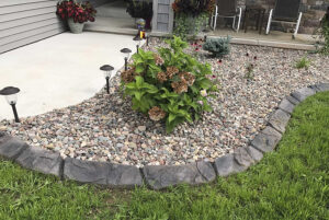A landscaped garden bed with decorative rocks, a hydrangea bush, solar lights, and a stone border creates a beautiful Showcase Gallery next to the concrete patio and grass lawn.