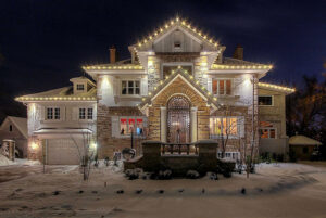 Large stone house at night, decorated with string lights along the roof, stands as a stunning showcase amid snow-covered ground and trees.
