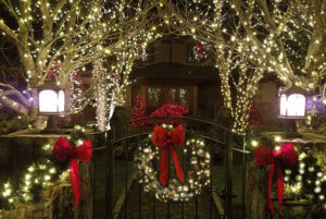 A house entrance becomes a festive showcase, adorned with holiday lights, a wreath on the gate, red bows, and illuminated trees and bushes glowing beautifully at night.
