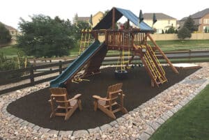 Showcase Gallery: Wooden playground set with slides, swings, and climbing features on mulch, surrounded by rocks, with two wooden chairs in the foreground and houses in the background.