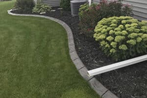A curved concrete garden edging defines a mulched flower bed with green shrubs from a neatly mowed lawn, creating an outdoor Showcase Gallery beside the house.