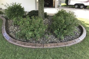 A landscaped garden bed with green shrubs and a tree, surrounded by decorative stone edging and filled with small rocks, creates a Showcase Gallery next to the lawn and driveway.