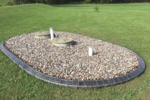A neatly bordered gravel area with two septic tank covers and two white vent pipes sits on a grassy lawn, creating a tidy Showcase Gallery for essential utilities.