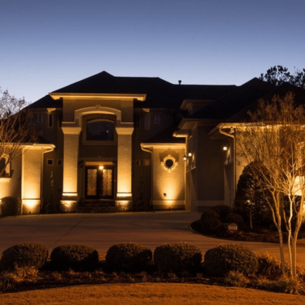 A large, modern Shorewood MN house with Holiday Lighting illuminating the entrance and driveway at dusk, surrounded by manicured shrubs, leafless trees, and elegant decorative concrete curbing.