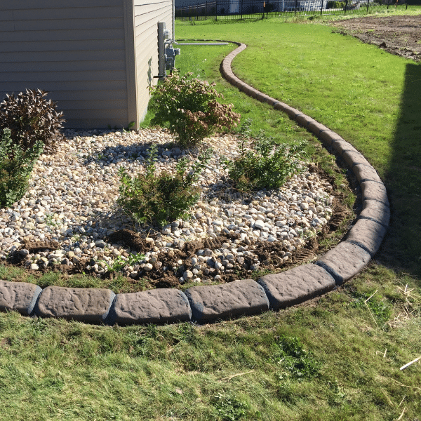 Landscaped garden bed with decorative rocks, concrete curbing, small shrubs, and grass, located beside a house in the Twin Cities and bordered by a curved pathway. Landscaped garden bed with decorative rocks, concrete curbing, small shrubs, and grass, located beside a house in the Twin Cities and bordered by a curved pathway.