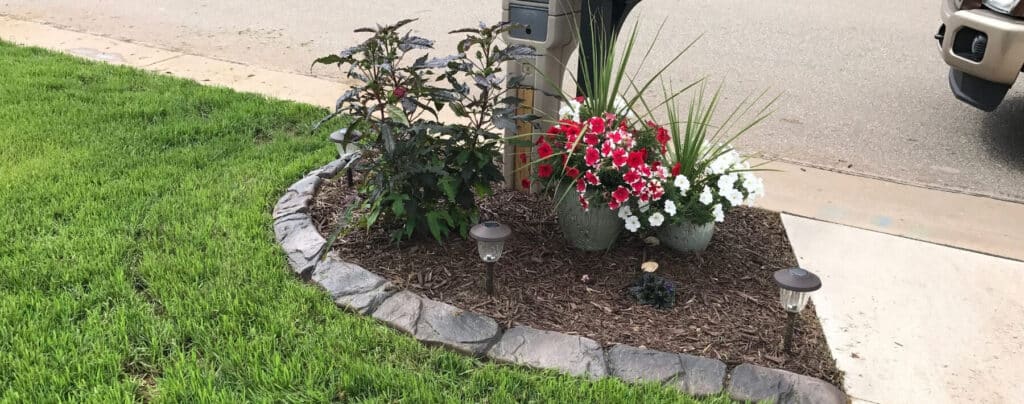 A small landscaped area with concrete curbing by the street features green grass, two potted flowering plants, decorative rocks, mulch, and solar garden lights near a mailbox.