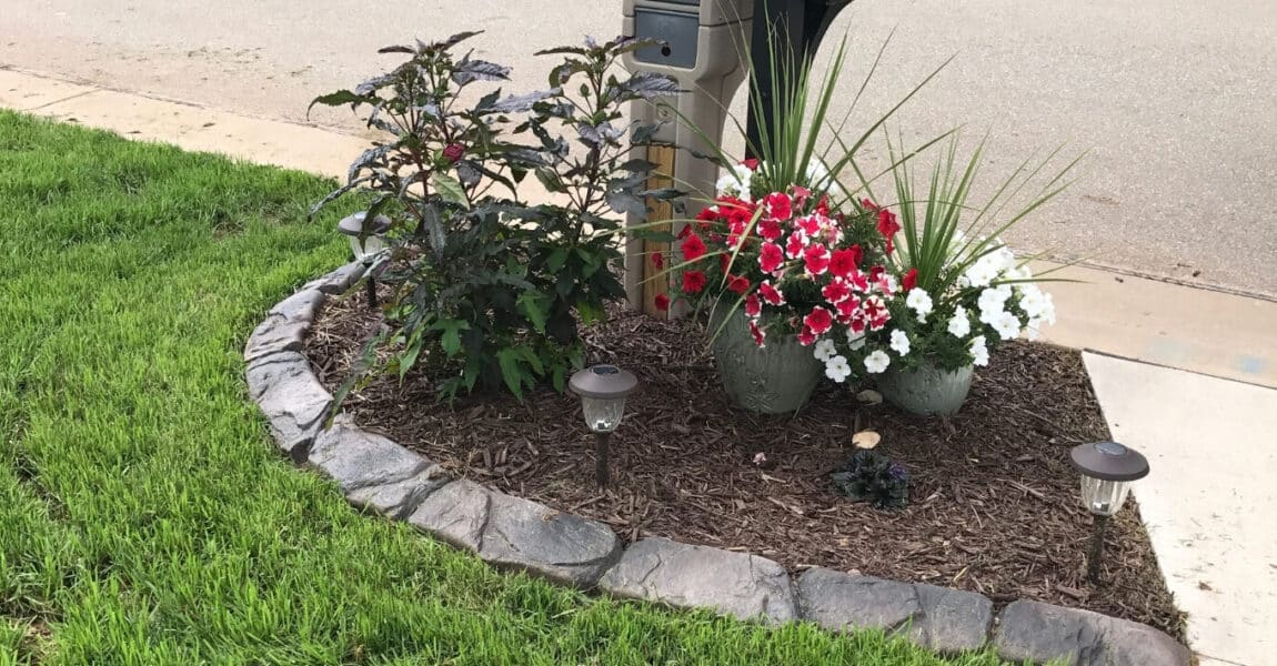 Concrete Curbing Basics 1 A small landscaped area with concrete curbing by the street features green grass, two potted flowering plants, decorative rocks, mulch, and solar garden lights near a mailbox.