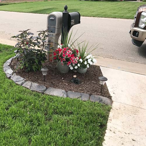A mailbox stands next to a small landscaped garden with flowering plants, mulch, and two solar lights, bordered by a stone edging near a driveway and lawn.