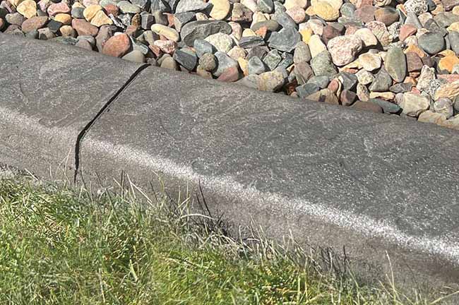 A concrete curb separates a patch of green grass from a bed of small, multicolored rocks.