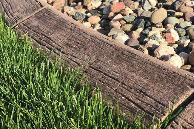 Close-up of a garden border with green grass on one side, a concrete edging designed to look like wood in the middle, and assorted rocks on the other side.